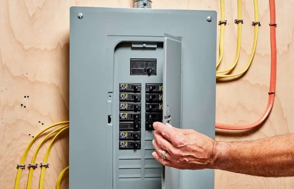 Hand opening the door of a home electrical panel with a main breaker and rows of circuit breakers