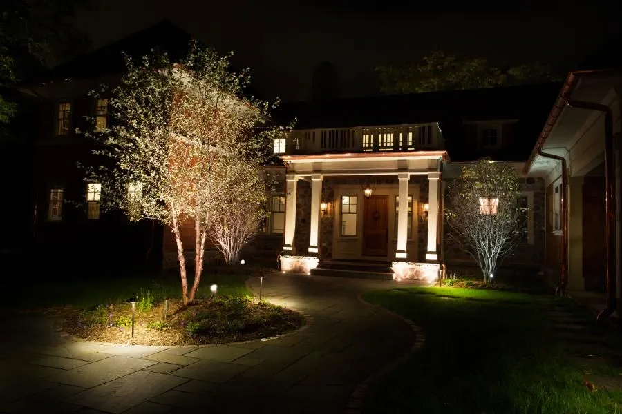 Front of a home at night with lit porch columns, uplit trees and path lights along a stone walkway