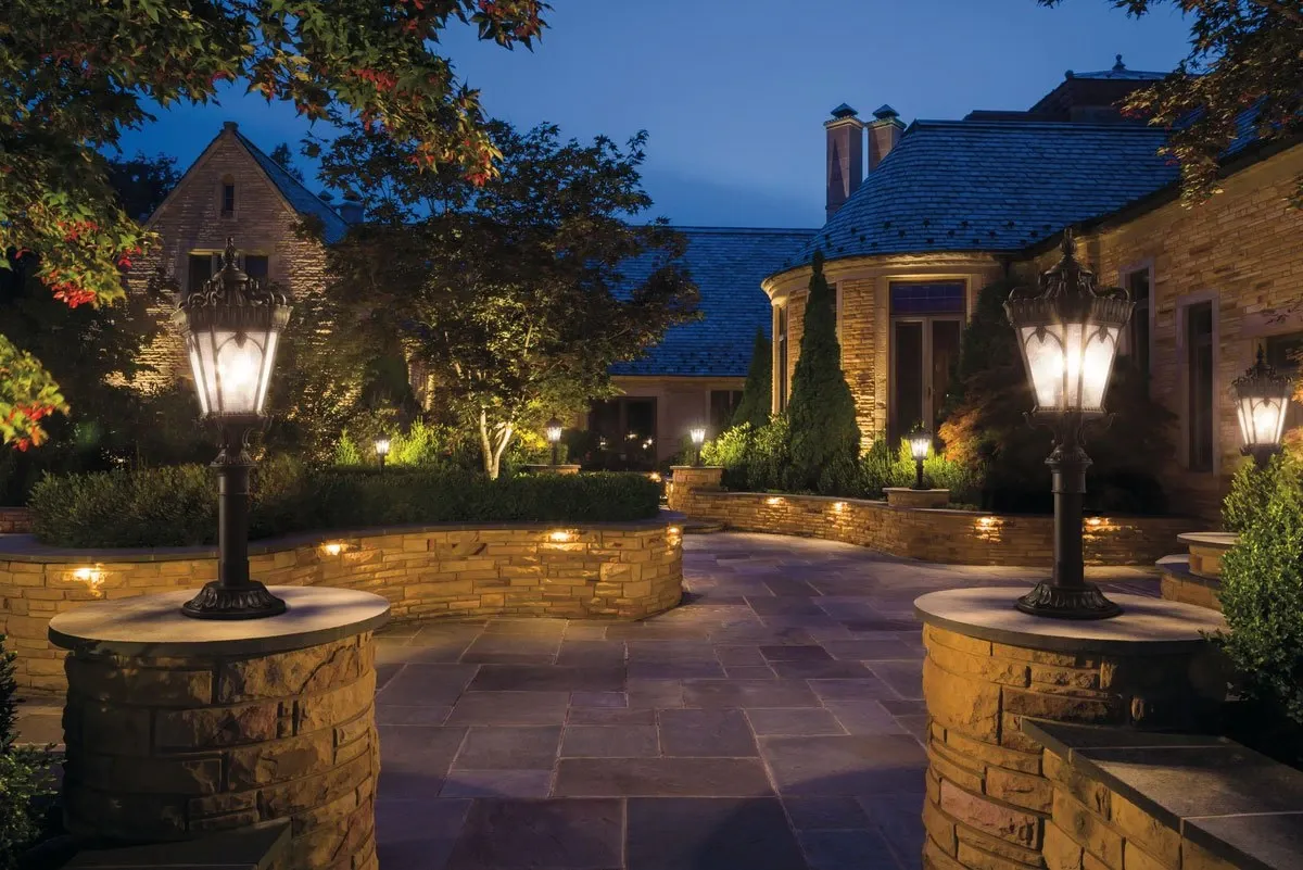 Stone patio at dusk lit by lantern post lights, step lights in the walls and uplights in the trees
